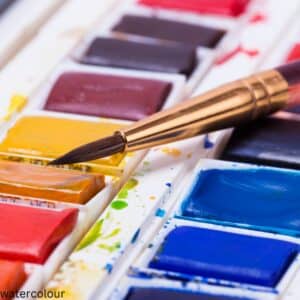 A close up of paintbrush resting on an open tin of various colours of watercolour paints.