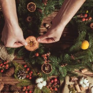 A person placing an ornament on a green Christmas wreath, surrounded by pinecones, berries and dried fruits.